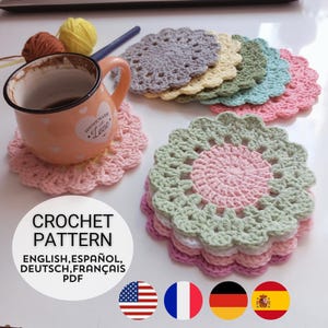 May include: A peach-colored ceramic mug with a heart design and the text "HOMEMADE Love" sits on a pink crochet coaster. Beside it are several round, scalloped-edge crochet coasters in pastel colors, yarn balls, a crochet hook, and a pattern label.