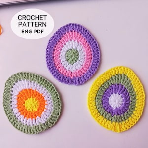 Puede incluir: Tres objetos coloridos en forma de huevo, hechos a ganchillo. Los huevos son en tonos verdes, naranjas, blancos, morados, rosas y amarillos. El texto "CROCHET PATTERN ENG PDF" está en un óvalo blanco en la esquina superior izquierda. Los huevos están sobre una superficie blanca.