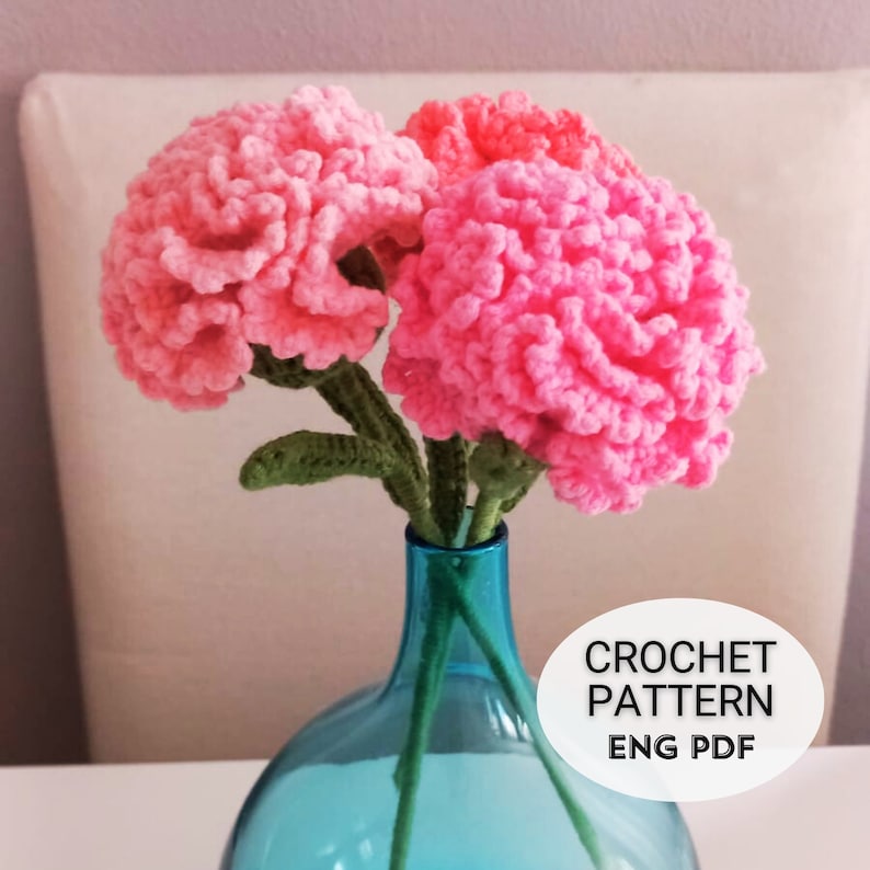 May include: Three pink crocheted carnations in a blue glass vase. The vase is on a white table. The image includes the text "CROCHET PATTERN ENG PDF".