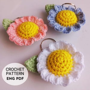 Op de afbeelding: Drie gebreide bloemen sleutelhangers in roze, blauw en wit met gele centra. Elke bloem heeft een groen blad. De sleutelhangers zijn bevestigd aan zilveren sleutelringen. De tekst "CROCHET PATTERN ENG PDF" is zichtbaar in de afbeelding.