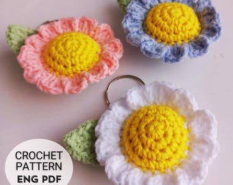 Könnte beinhalten: Drei gehäkelte Blumen-Schlüsselanhänger in Rosa, Blau und Weiß mit gelben Zentren. Jede Blume hat ein grünes Blatt. Die Schlüsselanhänger sind an silbernen Schlüsselringen befestigt. Der Text "CROCHET PATTERN ENG PDF" ist im Bild sichtbar.