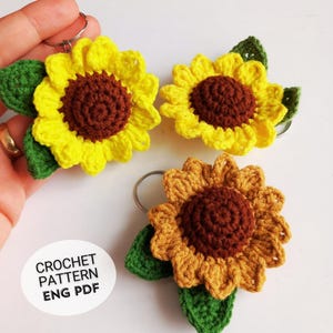 Puede incluir: Llaveros de girasol de ganchillo con pétalos amarillos, centros marrones y hojas verdes. Los llaveros están hechos a mano y presentan un diseño floral detallado. La imagen incluye el texto "CROCHET PATTERN ENG PDF".
