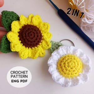 May include: Two crocheted flower keychains, one yellow sunflower with green leaves and one white daisy with a yellow center. The text "2 IN 1" is visible in the background. The text "CROCHET PATTERN ENG PDF" is visible in the foreground.