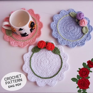 May include: Three crocheted coasters with floral embellishments. The coasters are in pink, blue, and white with green trim. The coasters are on a white surface. CROCHET PATTERN ENG PDF is written in black text on a white background.