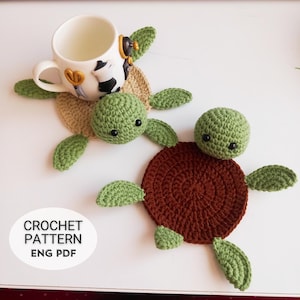 以下が含まれることがあります： 2つの亀の形をしたコースターのクロシェパターン。コースターは茶色とベージュの糸で作られており、黒い目の緑色の亀の頭が付いています。パターンは英語で入手可能で、PDFダウンロードです。