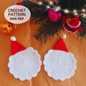 May include: Two white crocheted coasters with red crocheted Santa hats on top. The coasters are on a brown surface. The text "CROCHET PATTERN ENG PDF" is in the top left corner of the image.