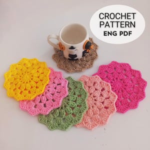 May include: Five crocheted coasters in different colors: yellow, pink, green, and peach. The coasters are round and have a flower-like design in the center. The text "CROCHET PATTERN ENG PDF" is visible in the top right corner of the image.