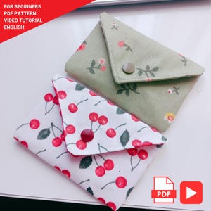 May include: Two handmade fabric pouches. One is white with a red cherry pattern, and the other is sage green with a floral design. Both have a button closure. The image includes text that says "FOR BEGINNERS PDF PATTERN VIDEO TUTORIAL ENGLISH".
