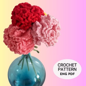 May include: A bouquet of crocheted carnations in shades of pink and red in a blue glass vase. The image includes the text "CROCHET PATTERN ENG PDF".