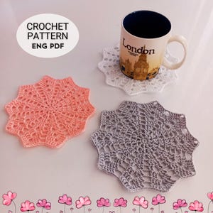 May include: Three handmade crochet coasters are visible. One is light pink, one is grey, and one is white. A mug with a London design sits on the white coaster. The image also includes the text "CROCHET PATTERN ENG PDF".