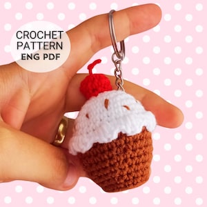 Könnte beinhalten: Häkelanleitung für einen Cupcake-Schlüsselanhänger mit einer roten Kirsche oben. Der Cupcake ist braun mit weißem Frosting. Die Anleitung ist in englischer PDF-Form verfügbar.
