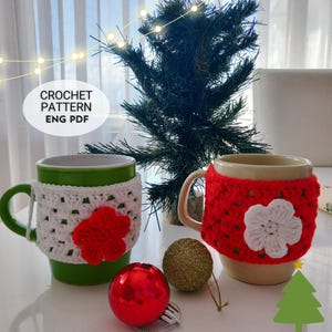 以下が含まれることがあります： 緑とベージュの2つのマグカップ。緑のマグカップには赤い花が付いた白いカバー、ベージュのマグカップには白い花が付いた赤いカバーが付いています。画像にはクリスマスオーナメントと小さなクリスマスツリーも含まれています。
