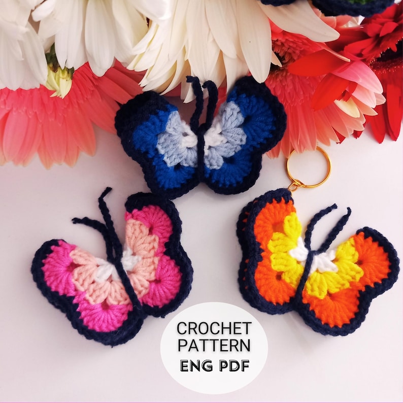May include: Three colorful crochet butterfly ornaments in shades of blue, pink, orange, and yellow. Each butterfly has a dark blue outline and delicate antennae. The text "CROCHET PATTERN ENG PDF" is visible. Flowers are in the background.