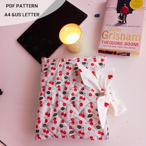 May include: A white quilted fabric with a cherry pattern, tied with a white bow featuring red hearts. A lit candle in a yellow cup, a book, and a black object are also present. Text on the image reads "PDF PATTERN A4 & US LETTER".