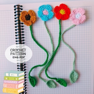 May include: Four handmade crochet flower bookmarks in orange, blue, red, and pink, each with a green stem and leaf. The text "CROCHET PATTERN ENG PDF" is in a white oval. The bookmarks are on a spiral notebook.