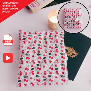 May include: A white quilted fabric with a red cherry pattern. A small, cherry-patterned book is tied with a white ribbon. A lit candle and a book are also visible. Text in the image reads: "FOR BEGINNERS PDF PATTERN VIDEO TUTORIAL ENGLISH."