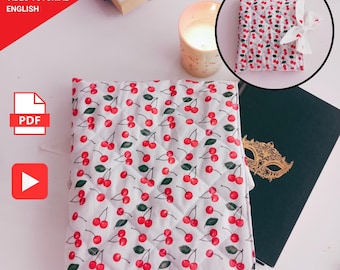 May include: A white quilted fabric with a red cherry pattern. A small, cherry-patterned book is tied with a white ribbon. A lit candle and a book are also visible. Text in the image reads: "FOR BEGINNERS PDF PATTERN VIDEO TUTORIAL ENGLISH."