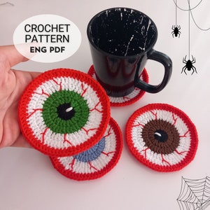 Pode incluir: Quatro tapetes de crochê em forma de olho, em vermelho, branco, verde, marrom e azul. Os tapetes estão sobre uma superfície branca com uma caneca preta e uma teia de aranha no fundo. O texto "CROCHET PATTERN ENG PDF" está no canto superior esquerdo da imagem.