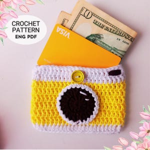 Op de afbeelding: Een handgemaakte geel en witte gehaakte camera-vormige portemonnee met een bruine lens en een gele knop. De portemonnee bevat een gele creditcard en Amerikaanse dollarbiljetten. De tekst "CROCHET PATTERN ENG PDF" staat in een witte cirkel.