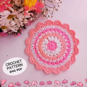May include: A crochet doily pattern in pink and white with a flower design in the center. The pattern is available as a digital download in English.