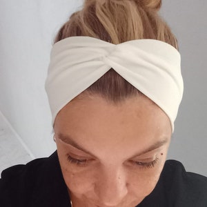 May include: A white turban headband with a twisted knot on top. The headband is made of a soft, stretchy fabric.