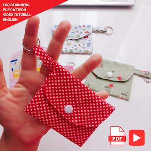 May include: A red polka dot fabric keychain pouch with a white button closure. The pouch is held up by a silver keyring. Other pouches with floral and patterned designs are in the background. Text in the image reads "FOR BEGINNERS PDF PATTERN VIDEO TUTORIAL ENGLISH".