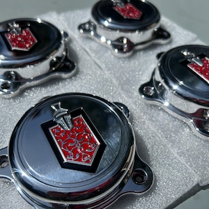 May include: Four chrome car centre caps. Each cap has a red and black emblem with a decorative design. The caps have a shiny, reflective surface and a three-hole mounting design.