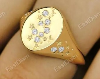 Puede incluir: Un anillo de sello dorado con un diseño celestial. El anillo tiene un acabado pulido y está adornado con pequeños diamantes, estrellas y una luna creciente. El anillo se muestra en un dedo.