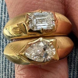 Peut inclure: Deux bagues en or avec de grands diamants clairs. L'une des bagues présente un diamant taille émeraude, l'autre un diamant en forme de poire. Les bagues ont une finition en or brossé et sont portées sur un doigt.