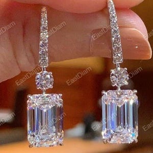 Könnte beinhalten: Elegante Ohrhänger mit klaren, smaragdgeschliffenen Edelsteinen. Die Ohrringe haben ein lineares Design mit kleinen, funkelnden Steinen, die zu einem größeren, rechteckigen Edelstein führen. Ein klassisches Schmuckstück.