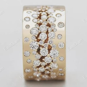 May include: A broad, gold-toned ring adorned with numerous sparkling, round diamonds of different sizes. The diamonds are arranged across the ring's surface, forming a textured, captivating pattern. The ring is displayed against a simple backdrop.