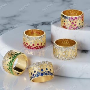 May include: A collection of gold rings with various gemstone embellishments. Some rings feature a gradient of red, blue, and green gemstones, while others are adorned with clear gemstones. The rings are displayed on a white marble surface.