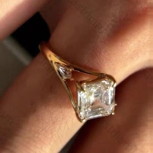 May include: A gold ring with a large, square-cut diamond as the focal point. The ring has a traditional design with a gold band and smaller diamonds flanking the main stone. The ring is set against a blurred background.