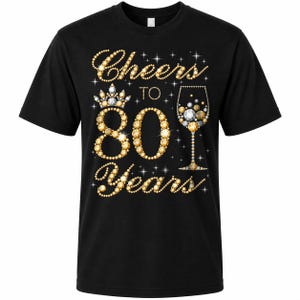 May include: A black short-sleeve t-shirt featuring a graphic design made of gold and silver faux-diamond beads. The text reads Cheers to 80 Years, accompanied by a crown icon and a wine glass filled with sparkling gold and silver spheres.