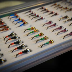 May include: A white plastic fly fishing box filled with rows of colourful flies. The flies are arranged in rows and are of various sizes and colours.