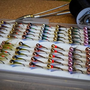 May include: A collection of colorful fishing flies arranged in rows on a white tray. The flies feature various colors, including gold, red, green, and pink, with metallic beads. A spool of thread and tools are in the background.