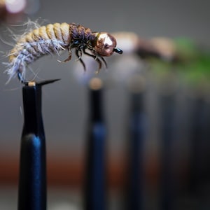 Może przedstawiać: Zbliżenie brązowo-beżowej przynęty muchowej z miedzianą główką. Przynęta jest zamontowana na czarnym stojaku.