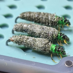 Könnte beinhalten: Vier grün-schwarze Fliegenfischerköder mit braunen und grauen, flauschigen Körpern.