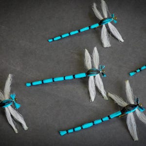 May include: Handmade dragonfly ornaments with turquoise and black segmented bodies and white wings. These decorative dragonflies are arranged on a dark grey surface, showcasing their delicate details and craftsmanship.
