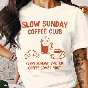 Può includere: T-shirt color crema con la scritta "SLOW SUNDAY COFFEE CLUB" in lettere color ruggine. Sotto, illustrazioni di una caffettiera, un croissant e una tazza di caffè. È incluso anche il testo "EVERY SUNDAY, 7-10 AM COFFEE COMES FIRST".
