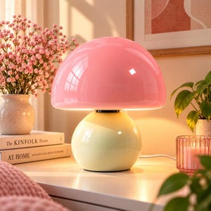 May include: A mushroom-shaped table lamp featuring a glossy, rounded pink shade and a smooth, pale yellow spherical base. The lamp emits a warm, soft glow from beneath the dome, creating a bright, illuminated surface on the base.