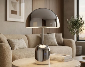 Puede incluir: Una lámpara de mesa con acabado en cromo pulido que presenta una gran pantalla redondeada en forma de cúpula y una base cilíndrica. La lámpara tiene un vástago central delgado que conecta la base con la pantalla y un pequeño botón circular.