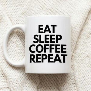 Könnte beinhalten: Ein weißer Keramikbecher mit einem Henkel auf der linken Seite. Auf der Vorderseite des Bechers befindet sich der schwarze Text EAT SLEEP COFFEE REPEAT in einer fetten, serifenlosen Schrift. Der Becher hat eine glatte Oberfläche.