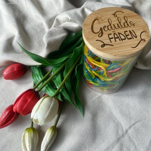 Könnte beinhalten: Ein klares Glas mit einem Holzdeckel, gefüllt mit bunten Gummibändern. Der Deckel ist mit dem Text "Gedulds Fäden" graviert. Künstliche rote und weiße Tulpen mit grünen Stielen und Blättern sind auf einem weißen Tuch angeordnet.