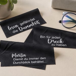 Könnte beinhalten: Drei rechteckige schwarze Mikrofasertücher mit gezackten Rändern. Jedes Tuch ist mit weißem Text in einer Mischung aus Schreib- und serifenloser Schrift bedruckt, darunter die Sätze "lesen gefährdet die Dummheit", "Bin für jeden Dreck zu haben" und "Martin Damit du immer den Durchblick behältst..." neben einer Brille mit schwarzem Rahmen.