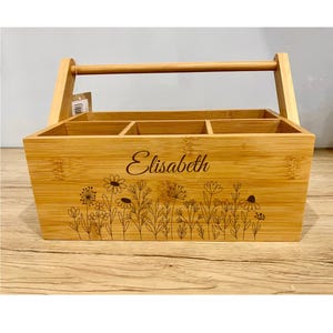 Könnte beinhalten: Ein Bambus-Utensilienhalter mit Griff und vier Fächern. Die Vorderseite des Halters ist mit dem Namen "Elisabeth" und einem Blumendesign graviert. Der Halter ist aus hellbraunem Bambus gefertigt.