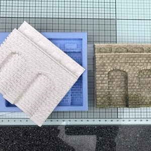Pode incluir: Um molde de silicone branco e um modelo de concreto cinza acabado de um arco de tijolo com duas aberturas. O molde está à esquerda e o modelo à direita.