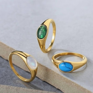 May include: Three gold rings, each with an oval gemstone. One has a green stone, another a white stone, and the third a blue stone. The rings are presented on a light grey surface.