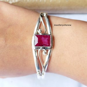 May include: A silver bangle bracelet with a delicate, ornate design features a large, rectangular, deep red ruby gemstone.