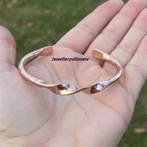 以下が含まれることがあります： ねじれたデザインのローズゴールドカラーのカフブレスレット。オープンカフは滑らかで光沢のある仕上げで、「Jewelleryvillanew」の文字が入っています。ブレスレットは手の上に表示されています。